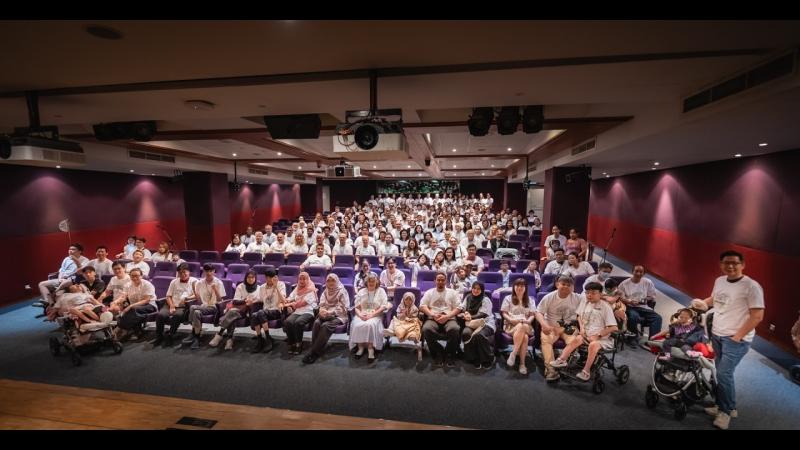 Group photo in an auditorium at the fifth Undiagnosed Hackathon in Singapore, September 2026: collaborators in white Undiagnosed Hackathon T-shirts fill the rows of purple seats, with the People Living With Undiagnosed Diseases and their families, several children in wheelchairs and strollers, in the front row.