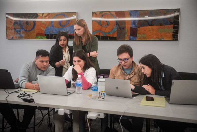 Six collaborators work on a case together at an Undiagnosed Hackathon: four sit at a table with laptops while two colleagues stand behind them, one pointing at a screen, in front of two large colourful paintings.